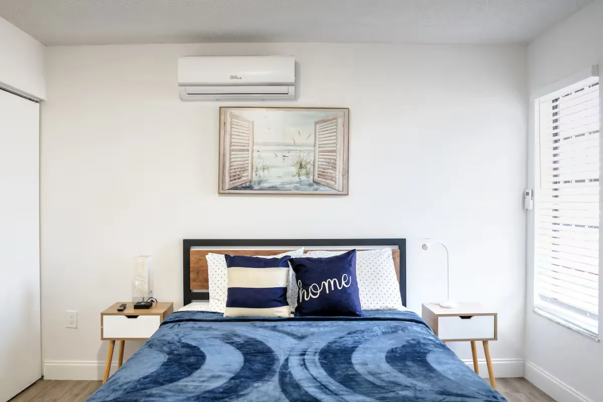 A cozy bedroom featuring a queen-size bed adorned with decorative pillows, artwork above the headboard, and a nightstand with a softly glowing lamp. Soft natural light fills the room, creating an inviting and restful atmosphere ideal for unwinding.
