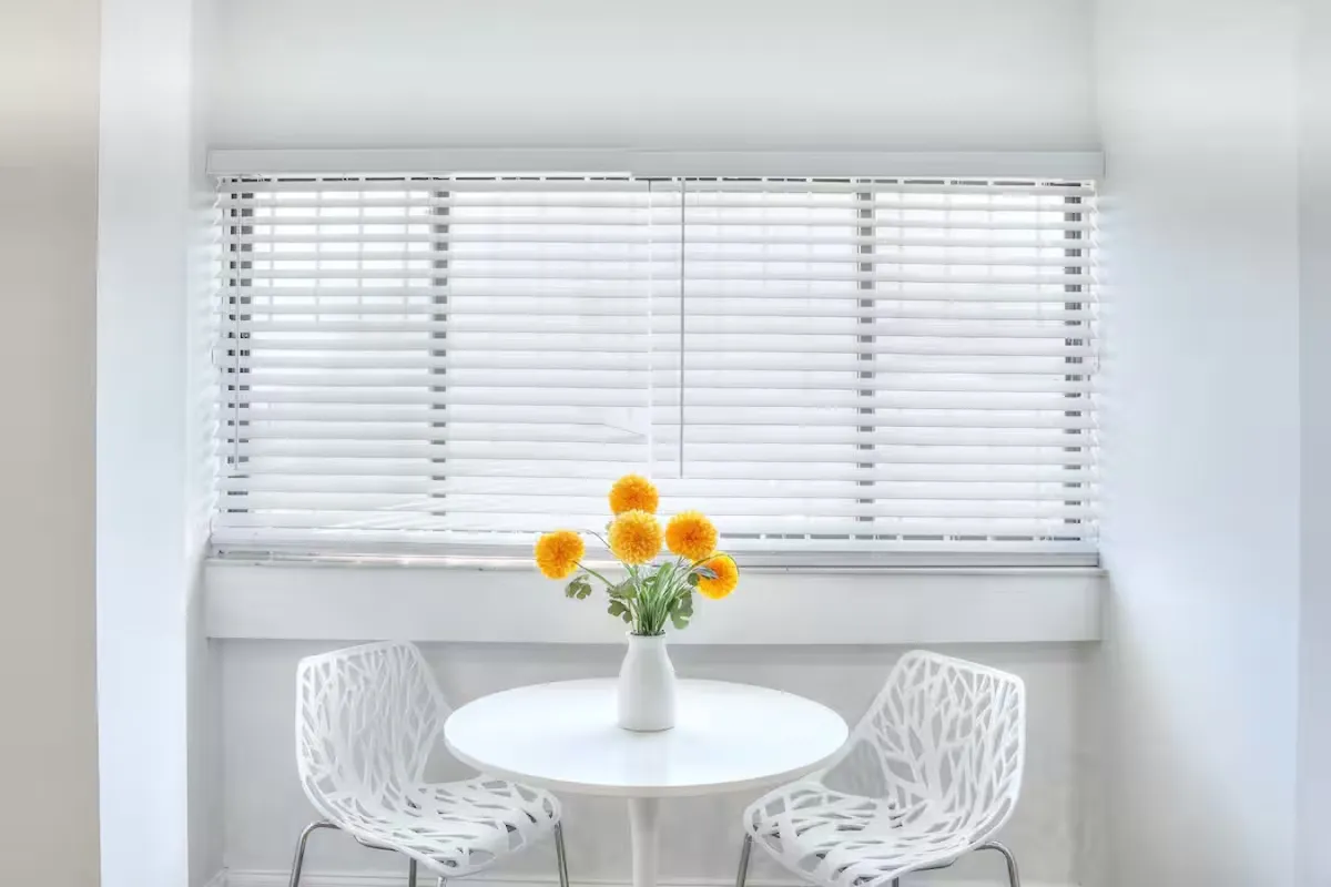 A cozy dining nook with a table adorned by a flower vase, surrounded by comfortable white chairs and illuminated by natural light streaming through window blinds. The space feels fresh and cheery, ideal for intimate meals or a quick breakfast.