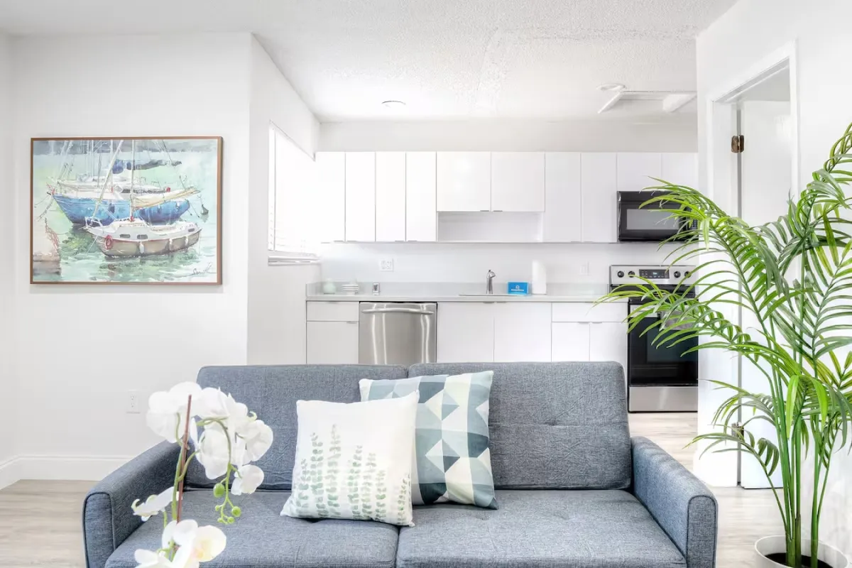 The image showcases a bright and airy open-plan living space featuring a modern gray sofa adorned with patterned pillows, a sleek white coffee table with decorative accents, and a fully equipped kitchen with white cabinetry and stainless steel appliances. A large framed mirror adds depth, and the setting is inviting for relaxation or socializing.