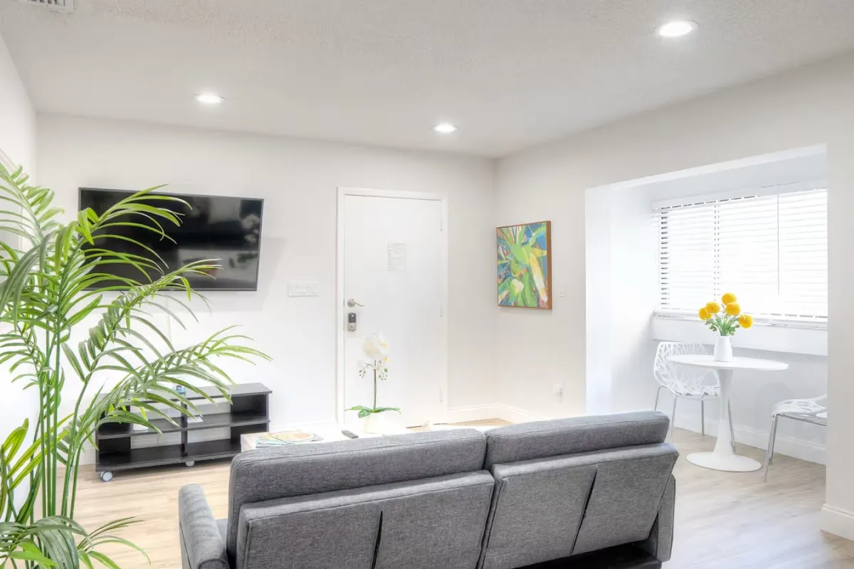 A bright, modern living room featuring a gray sofa with decorative pillows, a white coffee table topped with a magazine and plant, a large wall mirror with a white frame, and a view of the modern kitchen area with stainless steel appliances in the background. The open layout allows for seamless transition between relaxation and meal preparation.