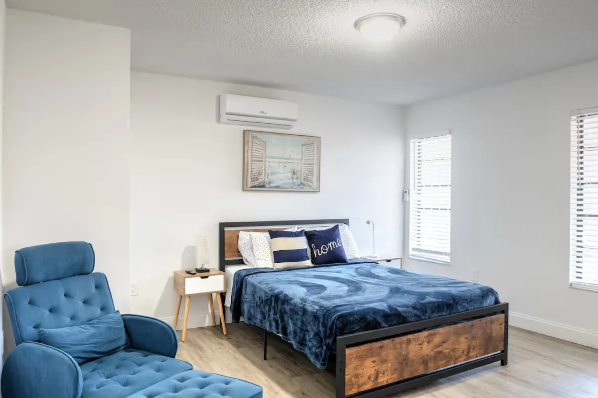 A cozy living room with a blue recliner chair and footrest, wooden cabinet under a wall-mounted TV, open doorway leading to the kitchen, and a blue fabric sofa. The scene suggests a comfortable setting for both relaxation and entertainment, with the kitchen easily accessible for meal prep.