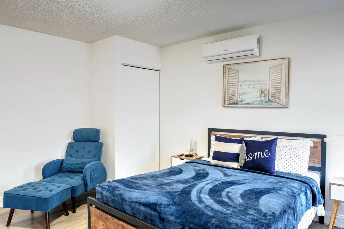 A serene bedroom featuring a queen-size bed with patterned blue bedding and decorative pillows, a blue upholstered chaise lounge chair with a matching footrest, framed seaside artwork above the bed, and a modern air conditioning unit mounted on the wall. Soft natural light and bedside lamps create a peaceful, inviting atmosphere.