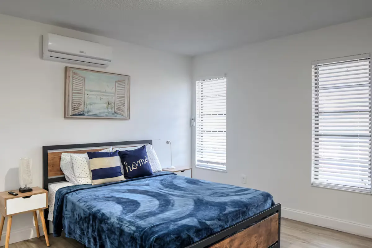 A bright, peaceful bedroom with a queen-size bed dressed in blue bedding and decorative pillows, wooden nightstands with subtle decor, an air conditioning unit above the bed, and three windows with white blinds that let in diffused natural light.