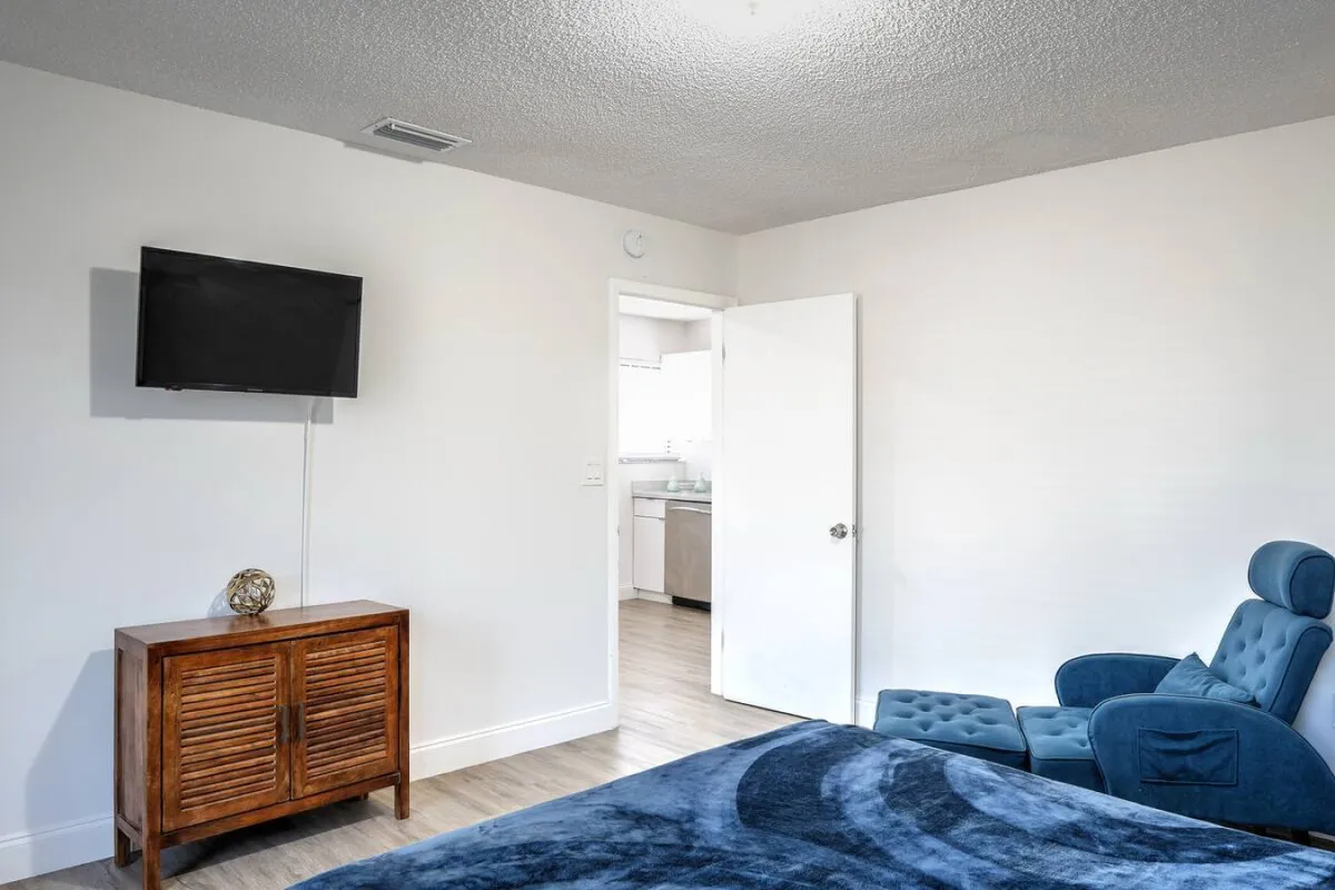 A tranquil living space featuring a blue reclining chair with matching ottoman, a wooden cabinet beneath a wall-mounted flat-screen TV, and an open doorway connecting to the kitchen. The space is bathed in natural light and designed for relaxation after a day out in the city.